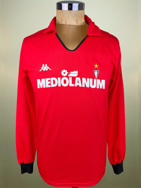 A red long-sleeve soccer jersey displayed on a mannequin torso. The jersey features a V-neck with black trim, black ribbed cuffs, and white printed logos including Kappa, the Mediolanum sponsor, and the AC Milan club crest.