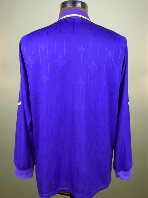 A purple long-sleeve soccer jersey with a subtle tonal fleur-de-lis pattern, displayed on a mannequin from the back. It features a yellow collar and yellow sleeve accents.