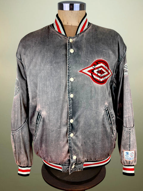 A vintage-style acid-wash denim varsity jacket displayed on a mannequin torso. The jacket features a faded gray denim body, a red, white, and black embroidered chest logo, and striped ribbed collar, hem, and cuffs.