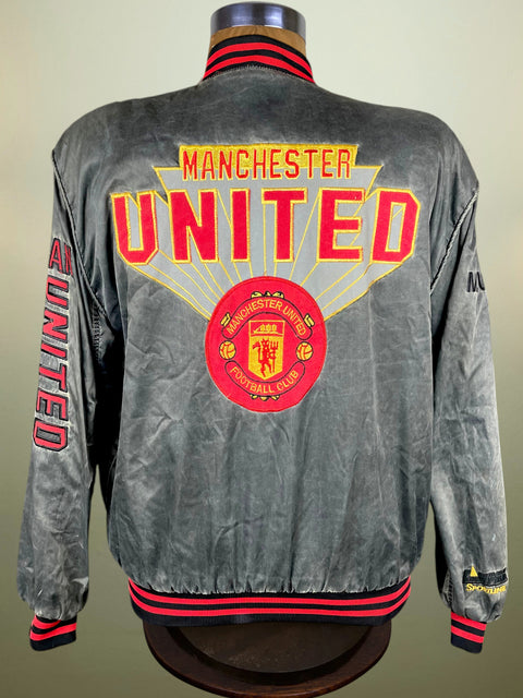A gray satin Manchester United bomber jacket displayed on a mannequin, showing the back view with 'MANCHESTER UNITED' in red and yellow, the club crest, and red and black striped ribbed collar, cuffs, and hem.
