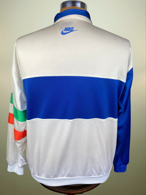 A vintage Nike track jacket shown from the back on a mannequin. The jacket features a white body with a wide royal blue horizontal stripe across the upper back, a blue Nike logo on the collar, and color-blocked details on the sleeves in green, white, and orange.