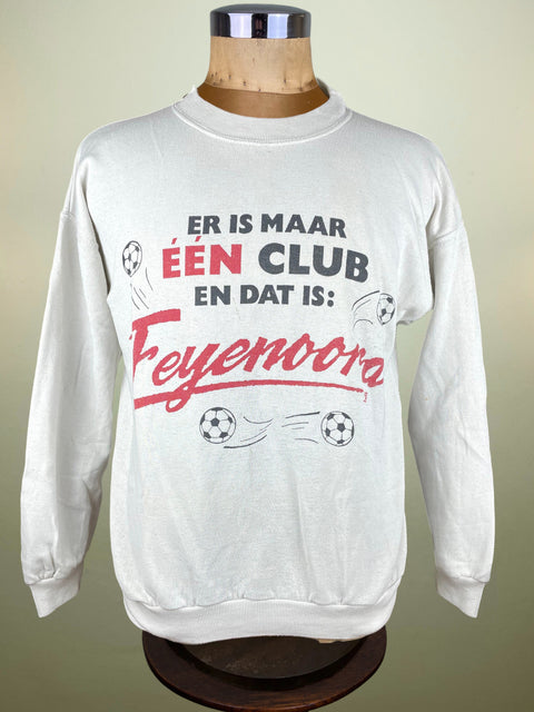 A white long-sleeve sweatshirt displayed on a wooden dress form mannequin. The sweatshirt features a graphic print in Dutch with the text 'ER IS MAAR ÉEN DAT IS: Feyenoora' in red and black lettering, accompanied by soccer ball illustrations.