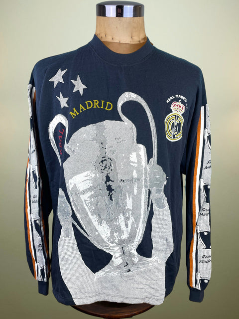 A navy blue long-sleeve Real Madrid soccer shirt with a large silver glittery Champions League trophy graphic on the front, displayed on a wooden mannequin torso against a neutral beige background.