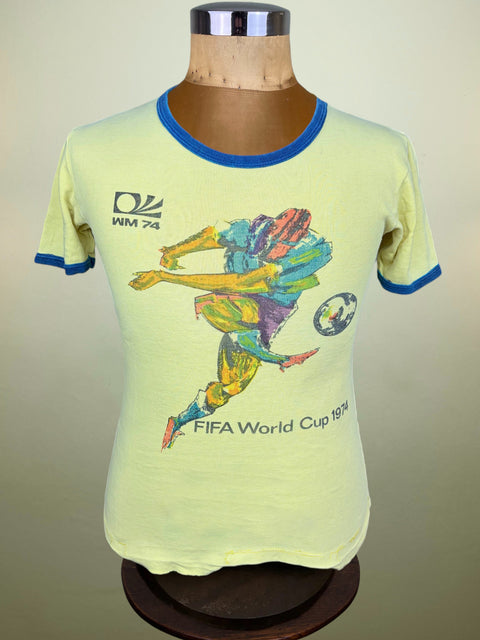 A vintage light yellow ringer t-shirt with blue trim, displayed on a wooden mannequin torso. The shirt features a colorful graphic of a soccer player kicking a ball, with 'FIFA World Cup 1974' and the WM 74 logo printed on the front.