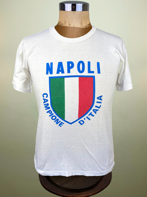 A white short-sleeve t-shirt displayed on a mannequin torso, featuring a blue 'NAPOLI' text graphic above a shield design with the Italian flag colors (green, white, red) and the words 'CAMPIONE D'ITALIA'.