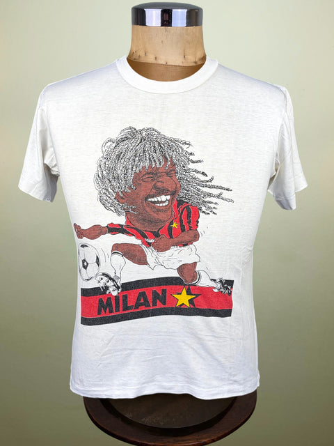 A white short-sleeve graphic t-shirt displayed on a wooden dress form mannequin. The graphic features a caricature of a football player with grey dreadlocks wearing a red and black striped jersey, kicking a white soccer ball, with a red banner below reading 'MILAN' and a yellow star.
