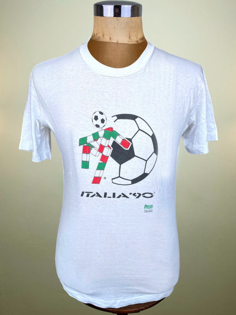 A white short-sleeve t-shirt displayed on a wooden mannequin torso, featuring a graphic of a soccer ball and a candy cane decorated with the Italian flag colors, with 'ITALIA'90' printed below.
