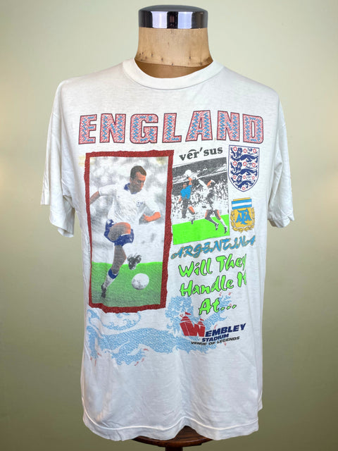 A vintage white England vs Argentina soccer t-shirt displayed on a wooden mannequin torso. The shirt features a large graphic print with the word 'ENGLAND' in red and blue patterned letters, a photo of a soccer player, the England crest, and text reading 'Argentina Will They Handle It Also'.