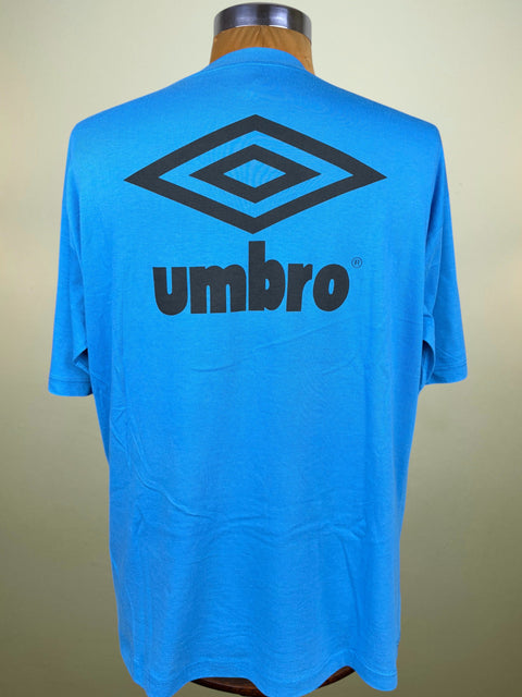 A bright blue short-sleeve Umbro t-shirt displayed on a wooden dress form mannequin, showing the back with a large black Umbro diamond logo and 'umbro' text.