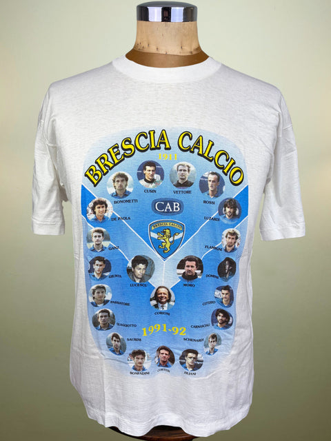 A white short-sleeve graphic T-shirt displayed on a wooden mannequin torso against a plain beige background. The shirt features a large circular blue print with the text 'BRESCIA CALCIO 1971-1980' and multiple player portraits arranged around a central CAB crest.