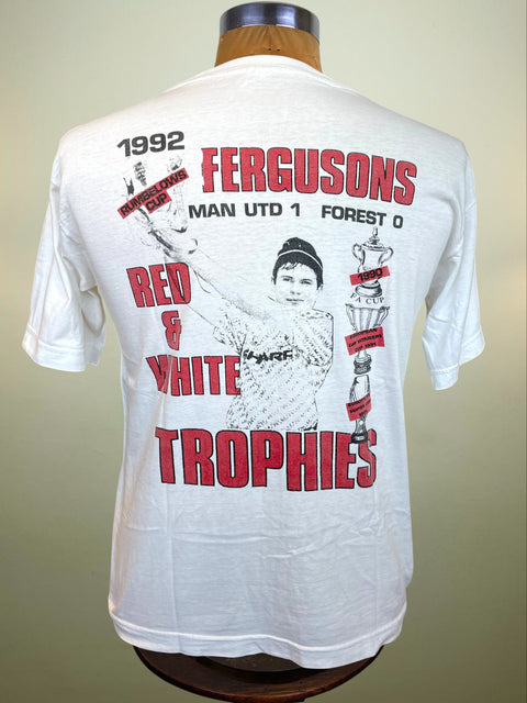 A white short-sleeve T-shirt displayed on a mannequin torso, showing the back side with a large red and black graphic print. The print reads '1992 FERGUSONS TROPHIES - MAN UTD 1 FOREST 0' and 'RED WHITE,' with a black-and-white illustration of a person holding trophies.