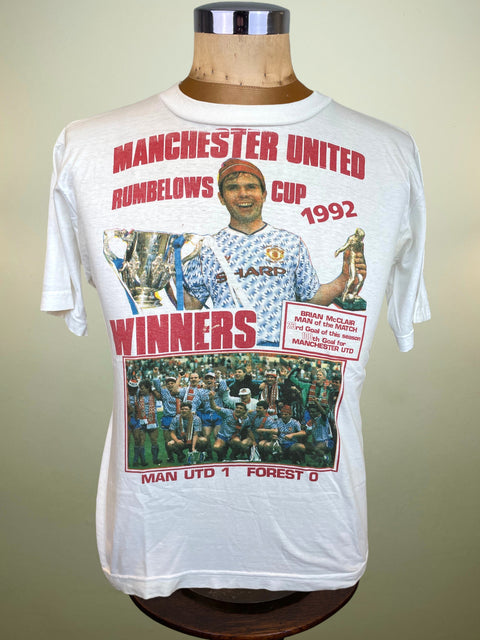 A white Manchester United 1992 Rumbelows Cup winners graphic t-shirt displayed on a wooden mannequin torso against a neutral beige background.