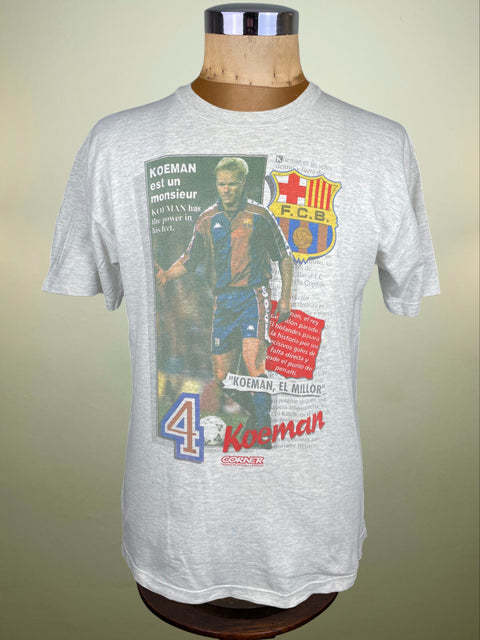 Gray t-shirt with a graphic of a soccer player and FC Barcelona logo on a mannequin against a beige background.