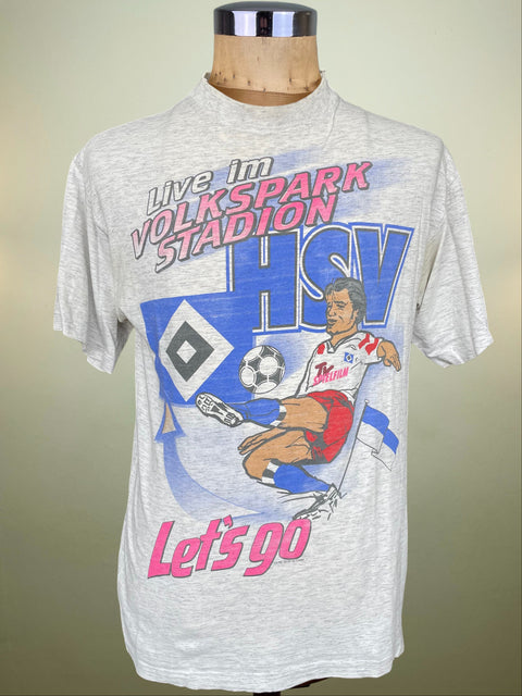 A light heather gray graphic t-shirt displayed on a wooden dress form mannequin. The shirt features a colorful print celebrating HSV with the text 'Live In VOLKSPARK STADION', a soccer player illustration, and 'Let's go' in pink lettering.
