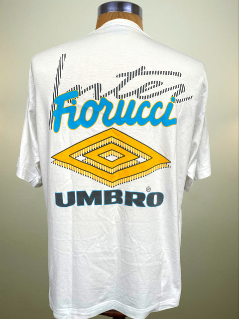 A white Umbro Fiorucci t-shirt displayed on a mannequin, showing the back graphic with a yellow diamond logo, blue 'Forucci' script, and dark blue 'UMBRO' text.