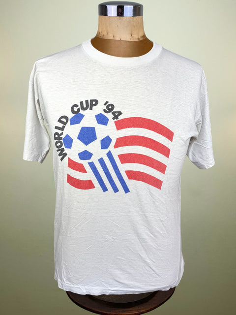 A white short-sleeve t-shirt displayed on a wooden dress form mannequin. The shirt features a graphic print of a soccer ball over a stylized American flag with the text 'WORLD CUP '94' arched above it.