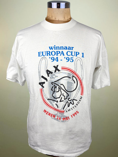 A white short-sleeve t-shirt displayed on a wooden dress form mannequin. The shirt features a graphic print celebrating Ajax Amsterdam's 1995 UEFA Champions League win, with blue, red, and black text and graphics.