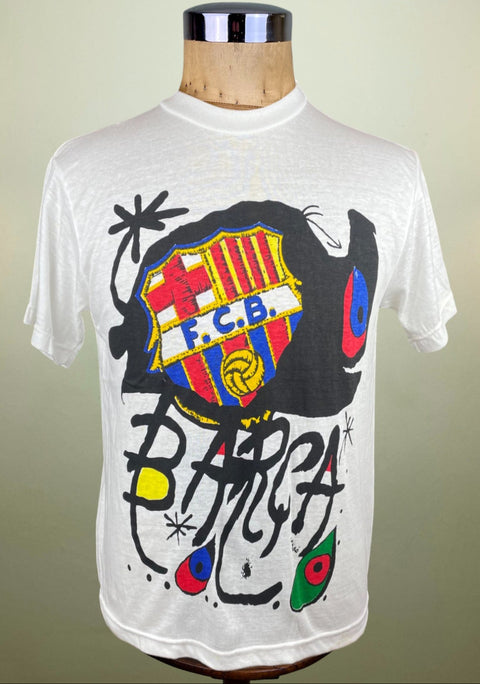 A white short-sleeve t-shirt displayed on a wooden mannequin torso, featuring a large graphic print of the FC Barcelona crest surrounded by black abstract graffiti-style lettering and colorful paint splatters.