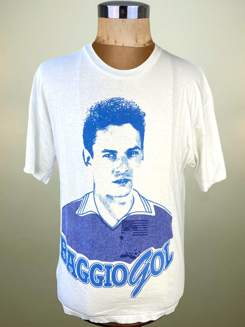 White t-shirt with blue graphic of a person and 'BAGGIO GOL' text on a mannequin against a beige background