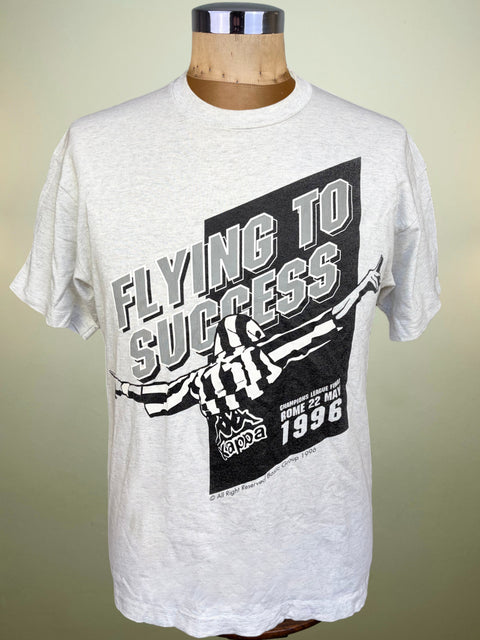 A light gray heather crewneck t-shirt displayed on a wooden dress form mannequin. The shirt features a large black and white graphic print with the text 'FLYING TO SUCCESS' and 'STOCKHOLM 1996'.