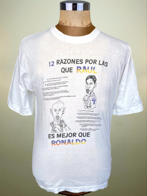 A white short-sleeve graphic t-shirt displayed on a wooden dress form mannequin. The shirt features a printed graphic with Spanish text reading '12 RAZONES POR LAS QUE RAUL ES MEJOR QUE RONALDO' and cartoon illustrations of both football players.