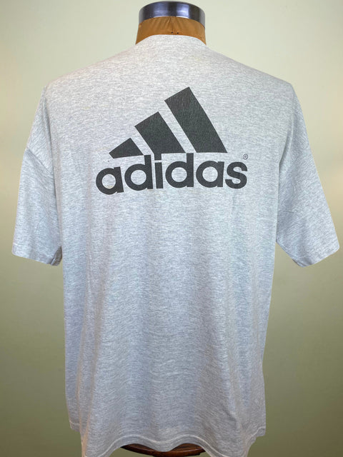 A light gray heather Adidas t-shirt displayed on a wooden dress form mannequin, showing the back with a large black Adidas logo and three stripes.