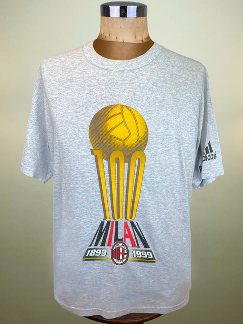 A light gray heathered Adidas t-shirt displayed on a wooden mannequin torso. The shirt features a large graphic of a yellow soccer ball on a trophy with '100', 'MILAN', '1899', and '1999' printed on it, and the Adidas logo on the right sleeve.