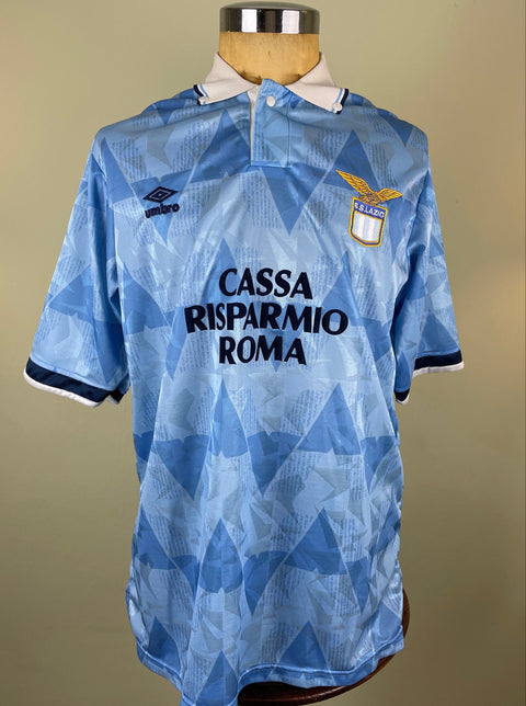 A short-sleeved Lazio 1990 home jersey featuring a geometric pattern in blue and white, with 'CASSA RISPARMIO ROMA' printed on the front and 'LAZIO' on the collar.
