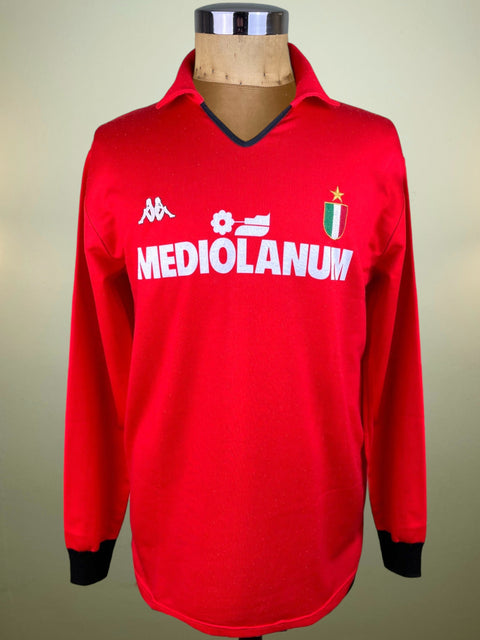 A red long-sleeve soccer jersey displayed on a mannequin torso. The jersey features a V-neck with black trim, black ribbed cuffs, and white 'MEDIOLANUM' sponsor text across the chest, along with the Kappa logo and the AC Milan club crest.