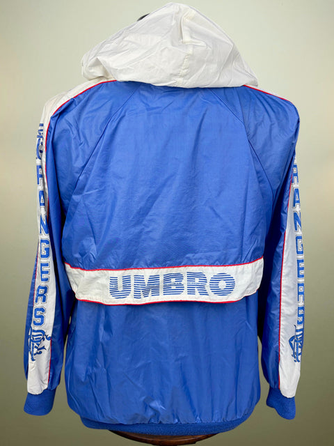 A blue and white Umbro windbreaker jacket shown from the back, featuring a white hood, a horizontal white stripe with 'UMBRO' in blue text, and vertical 'Rangers' text down the sleeves.
