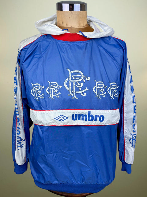 A blue Umbro windbreaker jacket displayed on a mannequin torso. The jacket features a white horizontal stripe across the chest with the blue Umbro logo, stylized white script across the back, and white sleeves with blue text.
