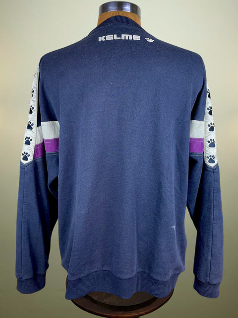 A navy blue long-sleeve sweatshirt with white and purple sleeve stripes and paw print details, displayed on a wooden dress form. The brand name 'KELME' is printed in white on the back collar.