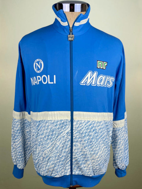 A blue and white Napoli soccer track jacket displayed on a mannequin. The jacket features a solid blue upper section with white trim, a patterned light blue and white lower section, and sponsor logos 'NAPOLI' and 'Mars' on the chest.