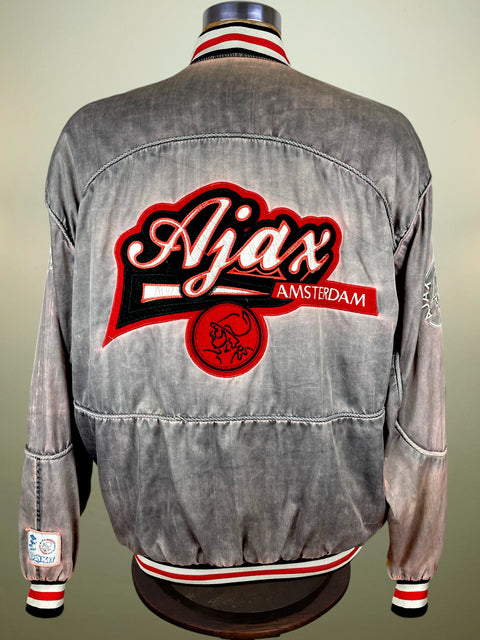 A gray acid-wash denim jacket shown from the back on a mannequin, featuring a large red and black 'Ajax Amsterdam' patch on the back with red and white striped ribbed collar, cuffs, and hem.