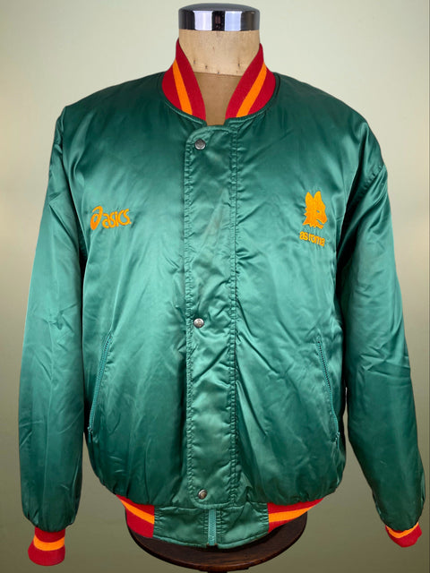 A green satin varsity-style jacket displayed on a dress form mannequin, featuring red and yellow ribbed trim on the collar, cuffs, and hem, with yellow embroidered AS Roma and Asics logos on the chest.