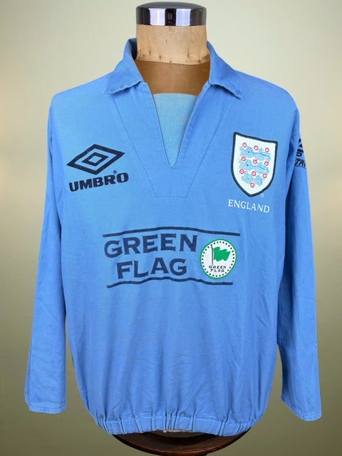 A light blue long-sleeve England soccer jersey displayed on a mannequin torso. The jersey features the Umbro logo on the right chest, the England crest on the left chest, and a large 'GREEN FLAG' sponsor logo across the center.