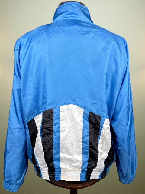 A blue windbreaker jacket shown from the back on a mannequin, featuring a high collar and a distinctive back panel with vertical black and white stripes bordered in blue.