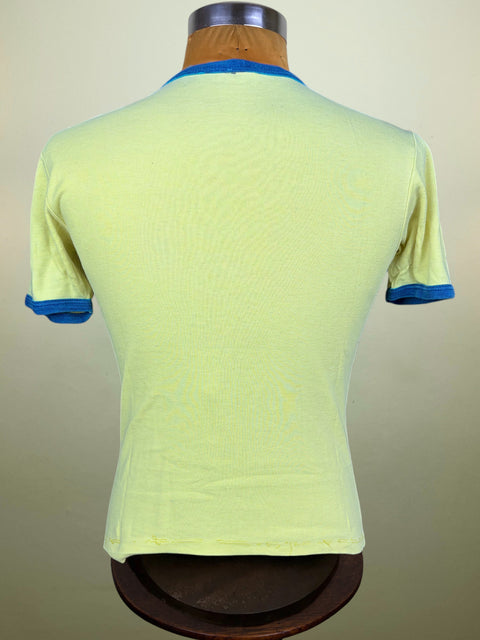 A light yellow-green short-sleeve t-shirt with blue ribbed trim on the collar and cuffs, displayed on a wooden dress form mannequin against a neutral beige background.