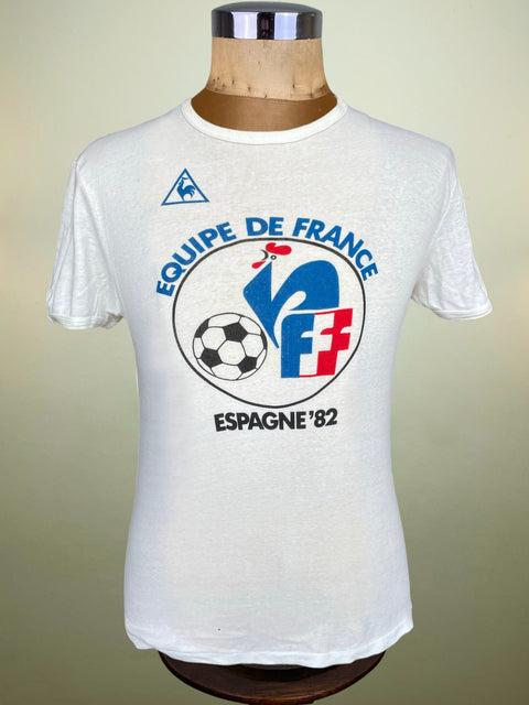 A white vintage short-sleeve t-shirt displayed on a wooden dress form mannequin. The shirt features a prominent blue, red, and black graphic for the 'Equipe de France' football team from the 1982 Spain championship, along with a small blue Le Malle logo on the upper chest.