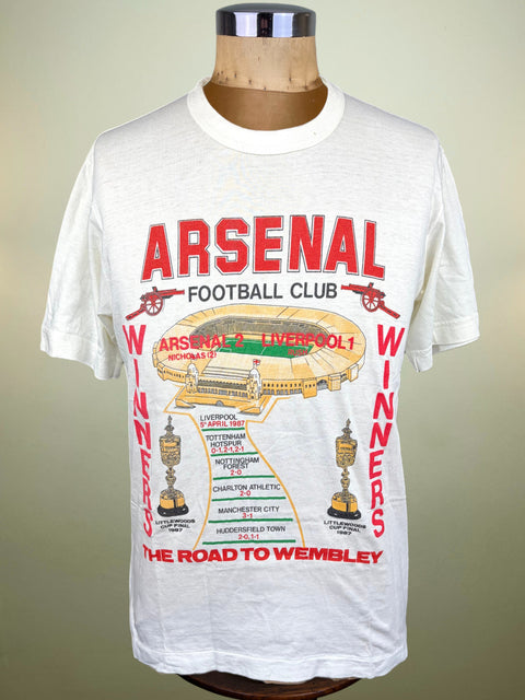 A white short-sleeve graphic T-shirt displayed on a mannequin torso. The shirt features a large red and multicolor print on the front reading 'ARSENAL FOOTBALL CLUB' with 'WINNERS' running vertically, plus a stadium illustration and trophy graphics.