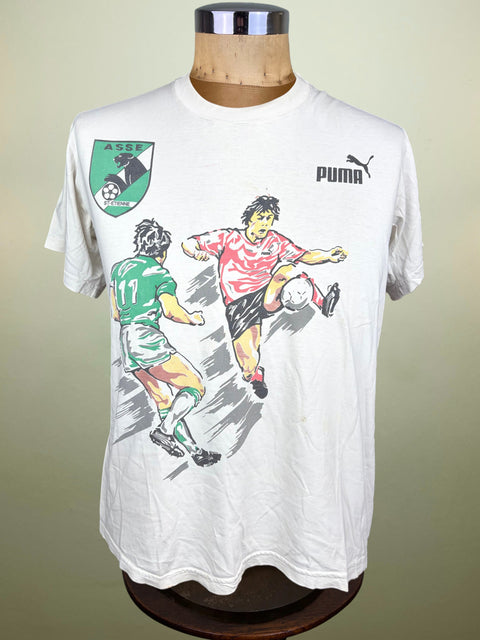A vintage white Puma soccer t-shirt displayed on a wooden mannequin torso. The shirt features a graphic print of two soccer players in green and pink/red jerseys, with the green AC Milan club crest on the left chest and the black Puma logo on the right chest.
