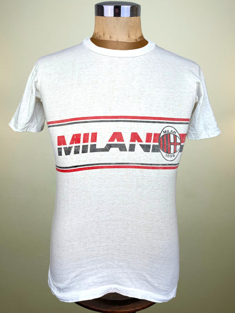A vintage white AC Milan t-shirt displayed on a wooden dress form mannequin. The shirt features a horizontal graphic stripe across the chest with red and black lines, the word 'MILANO' in red and black, and the AC Milan club crest on the right side.