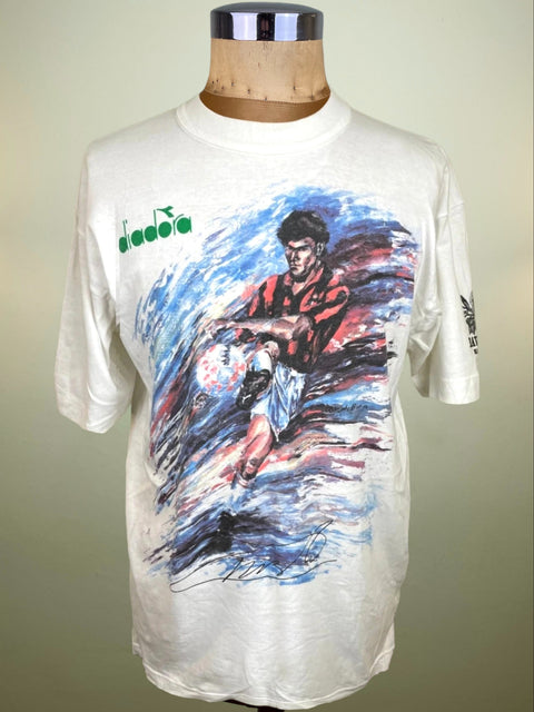 A vintage Diadora short-sleeve t-shirt displayed on a wooden dress form mannequin. The off-white t-shirt features a large graphic print of a soccer player in a red and black striped jersey, with blue and red brushstroke effects, and the green Diadora logo on the left chest.