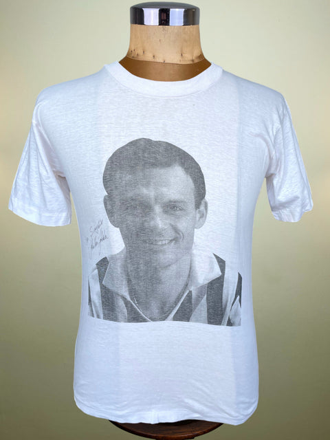 A white short-sleeve t-shirt displayed on a wooden mannequin torso against a beige gradient background. The shirt features a large, faded black-and-white graphic print of a smiling Toto Schillaci's face on the front.