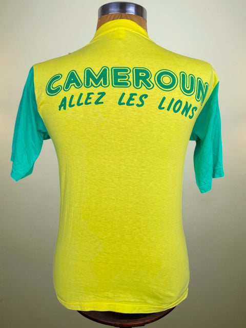 A short-sleeve T-shirt jersey displayed on a mannequin, showing the back view. The jersey is bright yellow with teal sleeves and green text reading 'CAMEROUN ALLEZ LES LIONS'.