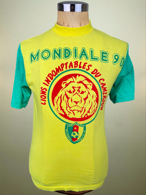 A vintage Cameroon football t-shirt displayed on a mannequin torso. The shirt features a bright neon yellow body with teal green short sleeves and a large red and green graphic on the front reading 'MONDIALE 90' and 'LIONS INDEMPTABLES DU CAMEROON' surrounding a lion emblem.