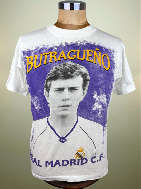 A white short-sleeve graphic T-shirt displayed on a wooden mannequin torso. The shirt features a large vintage-style print of footballer Butragueño with the name 'BUTRAGUEÑO' in yellow above and 'REAL MADRID'.