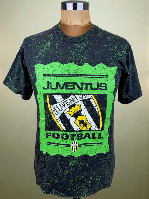 A vintage-style black t-shirt with a green splatter finish, featuring a distressed green graphic on the front with the text 'JUVENTUS FOOTBALL' and the Juventus club crest. The shirt is displayed on a wooden dress form mannequin against a neutral beige background.