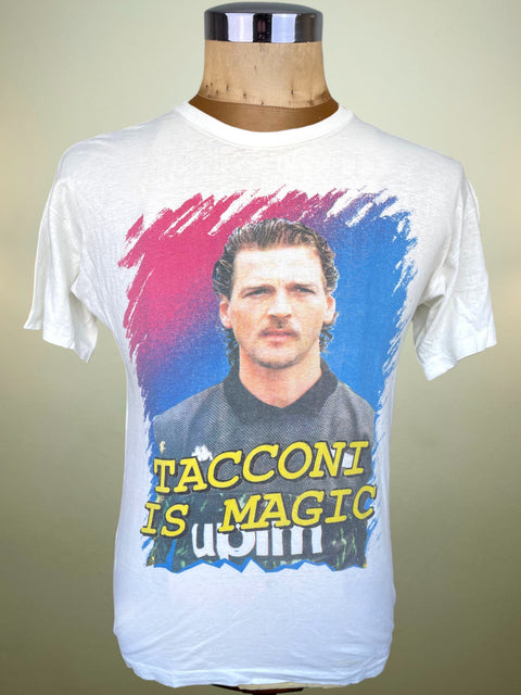 A white short-sleeve graphic t-shirt displayed on a wooden dress form mannequin. The shirt features a large front print of a man's portrait framed by red and blue paint strokes, with yellow text reading 'TACCONI IS MAGIC' and white text reading 'unli'.