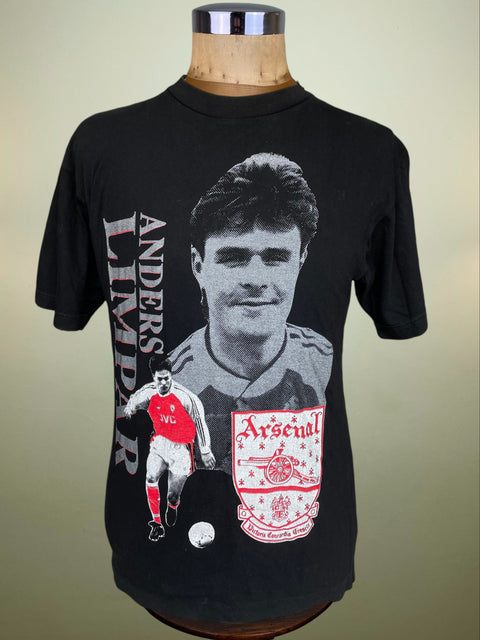 A vintage black short-sleeve t-shirt displayed on a wooden mannequin torso. The shirt features a large black-and-white graphic portrait of Anders Limpar, a smaller graphic of a player in a red and white Arsenal kit, a soccer ball, and the Arsenal crest on the front.